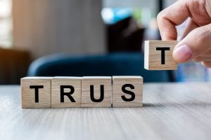 5 wooden blocks spell out the word "trust". A finger and thumb are shown to be placing the final letter in place. 
